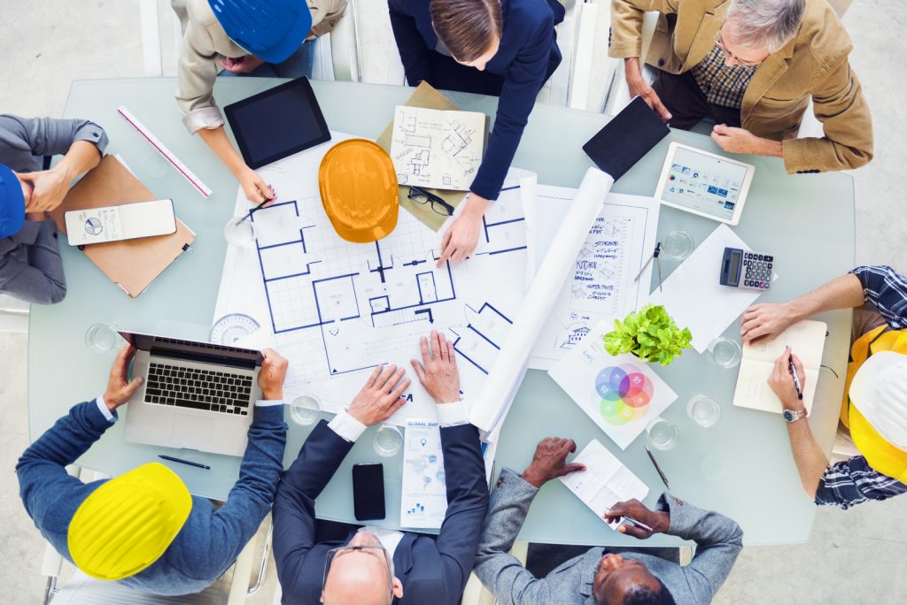 Lots of people in different construction roles working together at a table with plans, charts and devices 