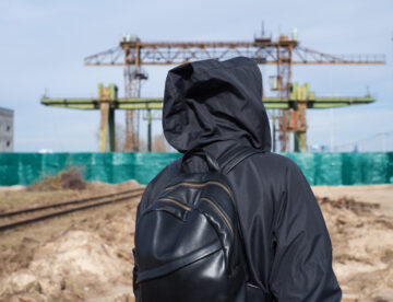 Back view of a man in dark clothing and carrying a bag with a construction site ahead of him