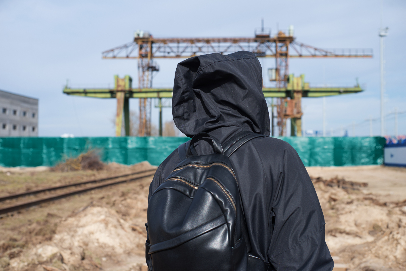 Back view of a man in dark clothing and carrying a bag with a construction site ahead of him