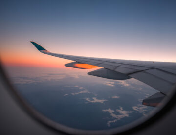 Image taken from an aeroplane showing the wing of the plane, the land below and a sunset in the distance