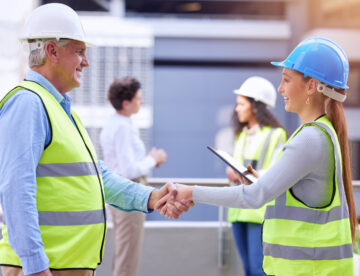 Image showing two construction professionals (a man and a woman) on a construction site shaking hands with other workers in the background.