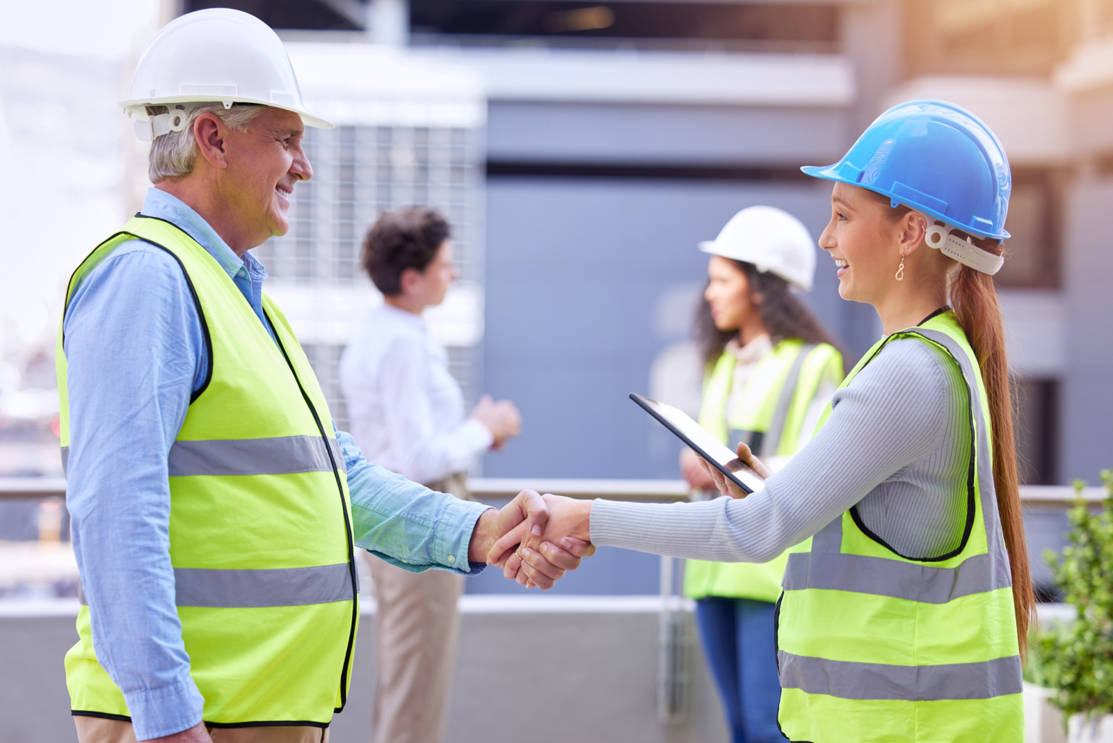 Image showing two construction professionals (a man and a woman) on a construction site shaking hands with other workers in the background.