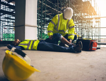 Image of an injured construction worker lying flat on the ground while another person attends to their leg.