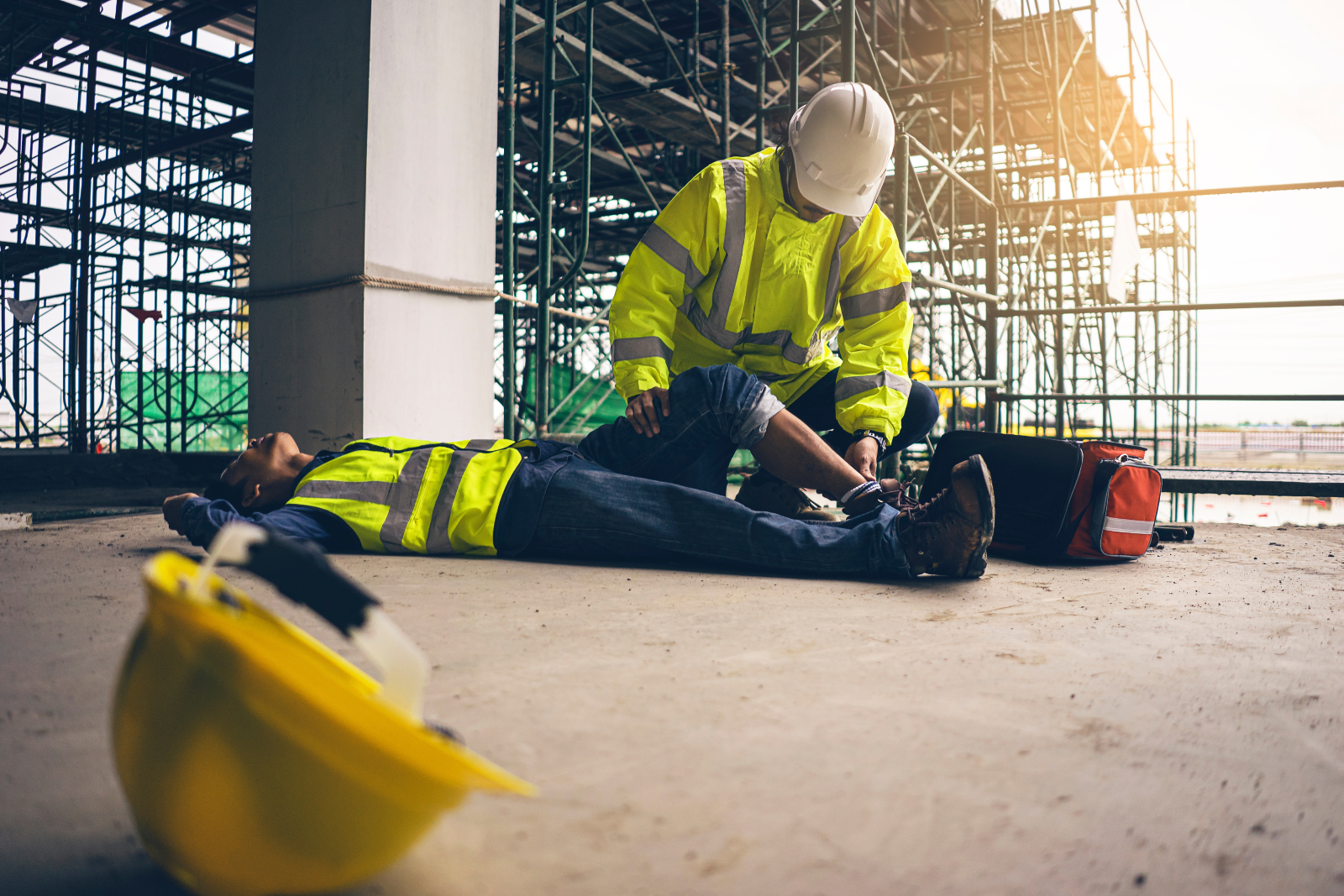 Image of an injured construction worker lying flat on the ground while another person attends to their leg.