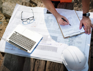 Image showing various items laid out on a table - construction plans, a laptop, a clipboard, glasses and a safety helmet