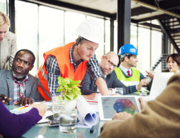 Image showing a diverse construction team working collaboratively around a table, some looking documents and IT equipment.