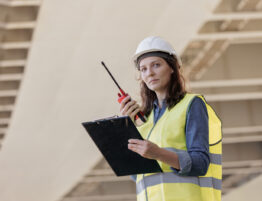 Woman wearing a high vis jacket and hard hat, holding a clipboard and a walkie-talkie with part of a construction site in the background