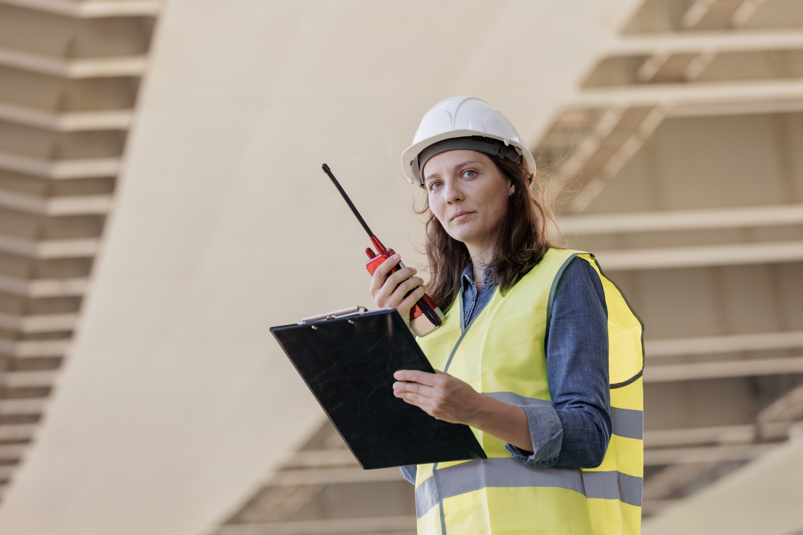 Woman wearing a high vis jacket and hard hat, holding a clipboard and a walkie-talkie with part of a construction site in the background