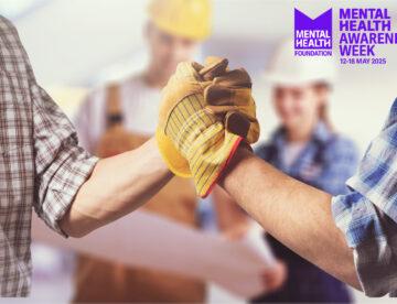 Close up of two construction workers arms (with hands shaking) with other workers standing behind them. Also featuring the Mental Health Awareness Week logo.
