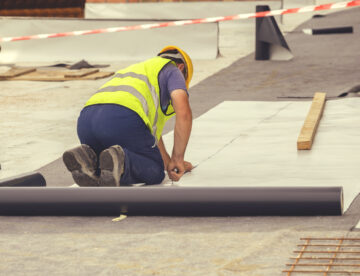A construction worker wearing a high-vis jacket, kneeling while laying a roofing system