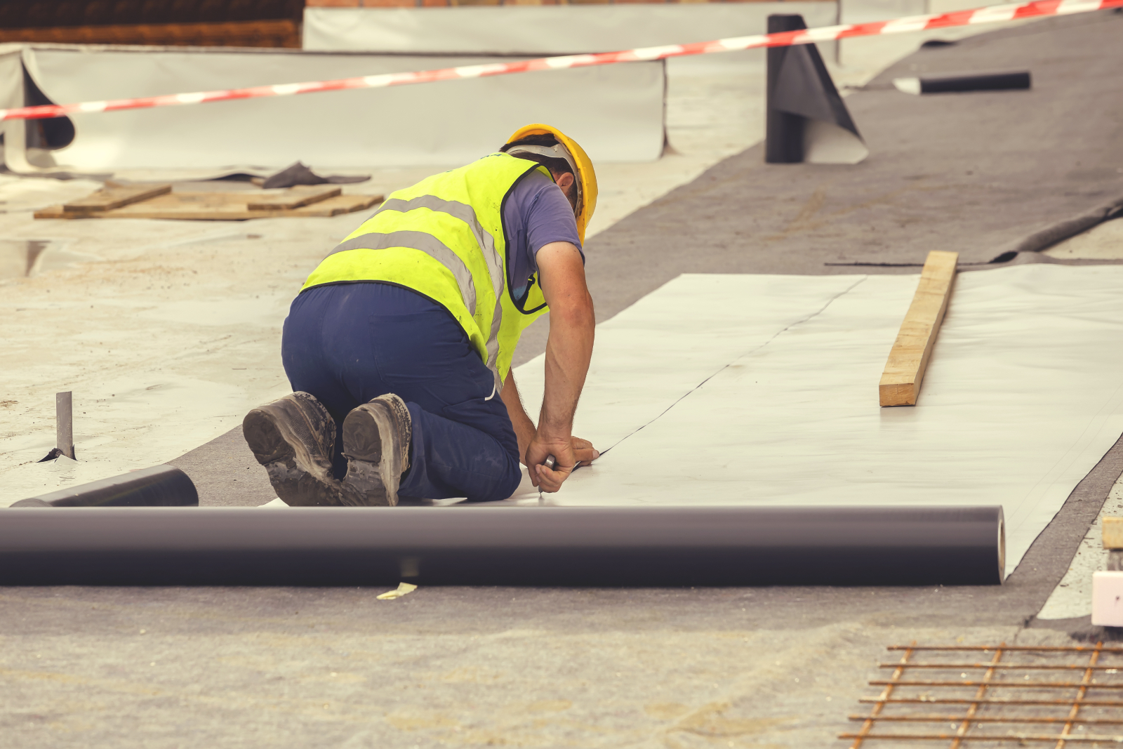 A construction worker wearing a high-vis jacket, kneeling while laying a roofing system