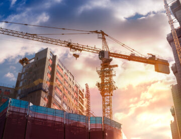 Construction site with a large building and a crane lifting materials, mix of clouds and sunshine in the sky behind the building