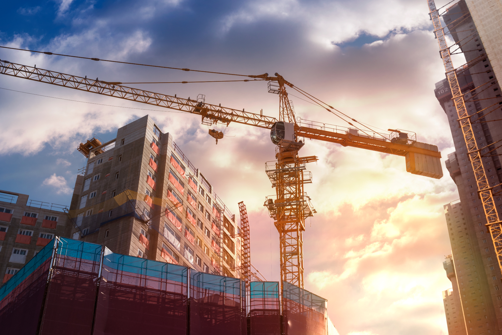 Construction site with a large building and a crane lifting materials, mix of clouds and sunshine in the sky behind the building