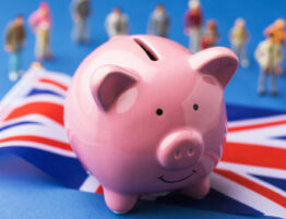 Close up of a pink piggybank sitting upon a UK flag and blurry toy plastic people behind it
