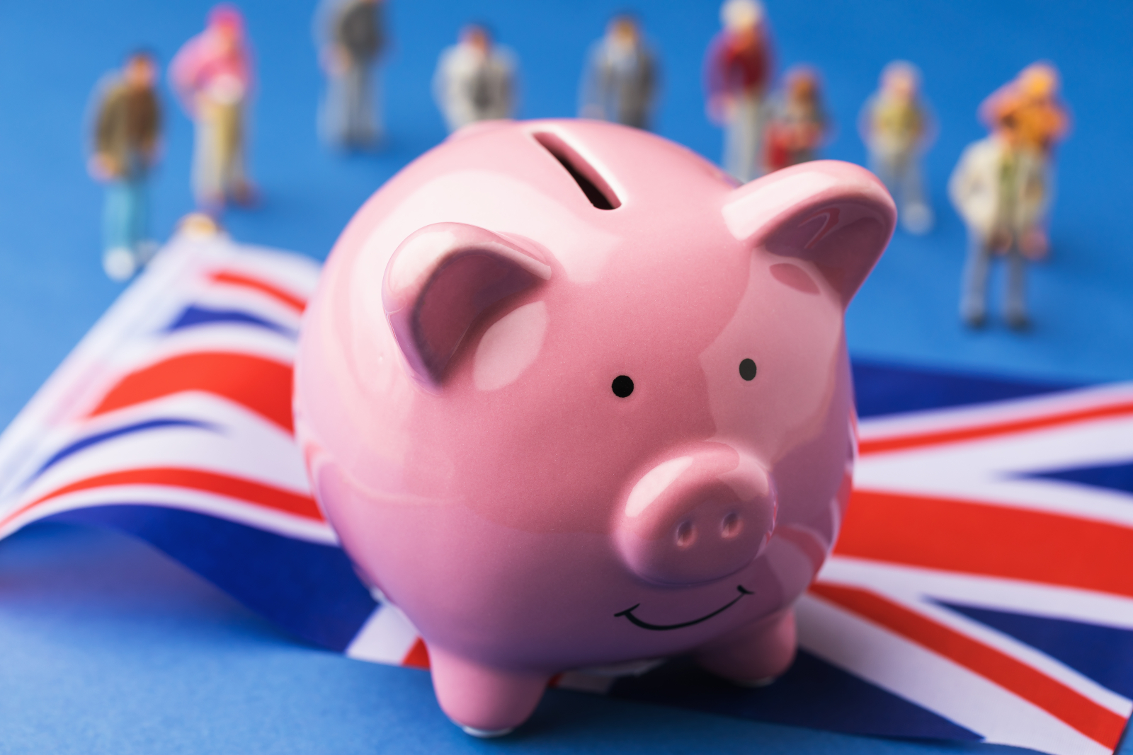 Close up of a pink piggybank sitting upon a UK flag and blurry toy plastic people behind it