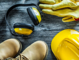 Selection of construction-related PPE set against a hard wooden floor. Items include a hard hat, boots, ear defenders and gloves.