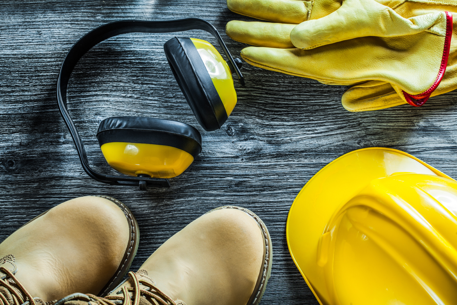 Selection of construction-related PPE set against a hard wooden floor. Items include a hard hat, boots, ear defenders and gloves.
