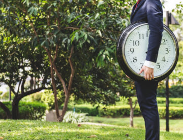 Image of a man standing in a park holding a giant clock. The man is on the right hand side of the image and only his torso and legs are visible. There is a large tree in the background to the left side of the image.