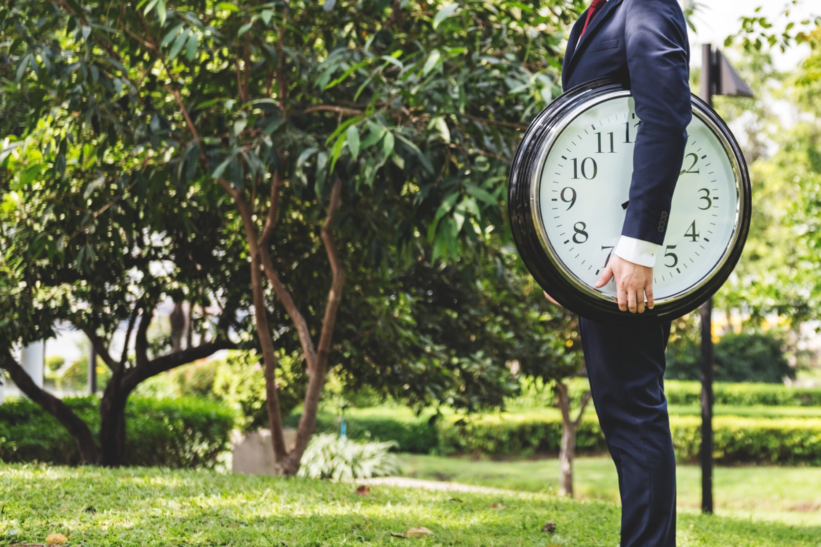 Image of a man standing in a park holding a giant clock. The man is on the right hand side of the image and only his torso and legs are visible. There is a large tree in the background to the left side of the image.