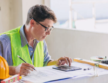 Construction manager in a high vis vest, sitting at an office desk looking over some paperwork and using a calculator