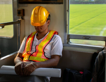 Construction worker sitting on public transport with his hard hat pulled over his face and shoulders slumped, indicating tiredness and possible stress