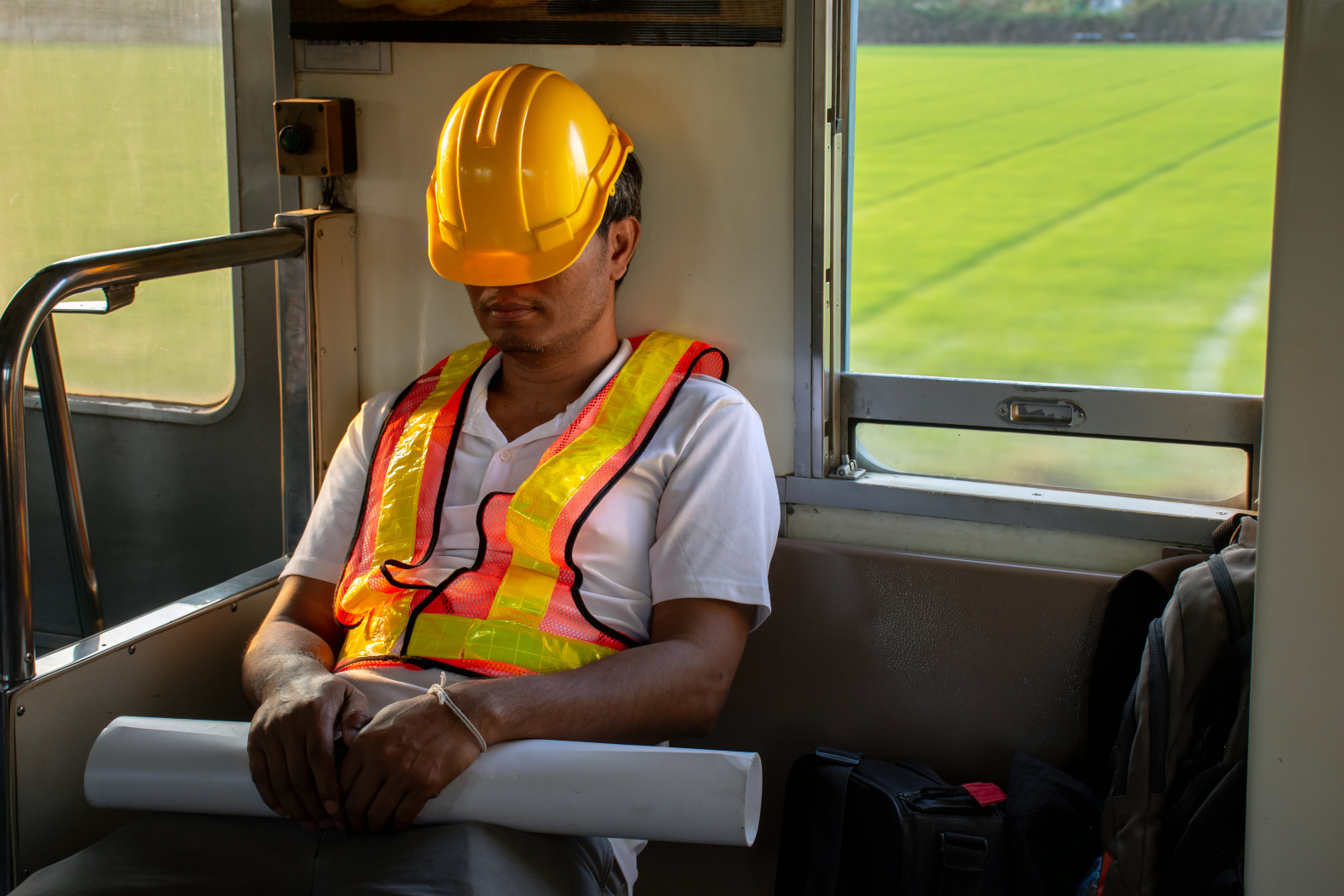 Construction worker sitting on public transport with his hard hat pulled over his face and shoulders slumped, indicating tiredness and possible stress