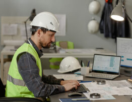 Construction worker sitting at a desk typing into a keyboard. There are papers, a hard hat and another laptop on the desk.