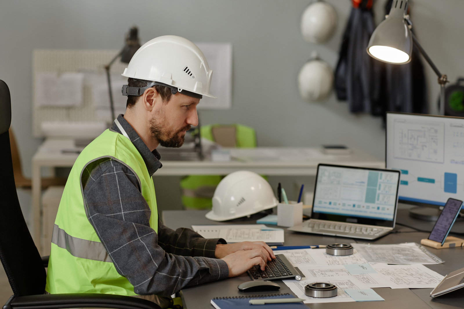 Construction worker sitting at a desk typing into a keyboard. There are papers, a hard hat and another laptop on the desk.