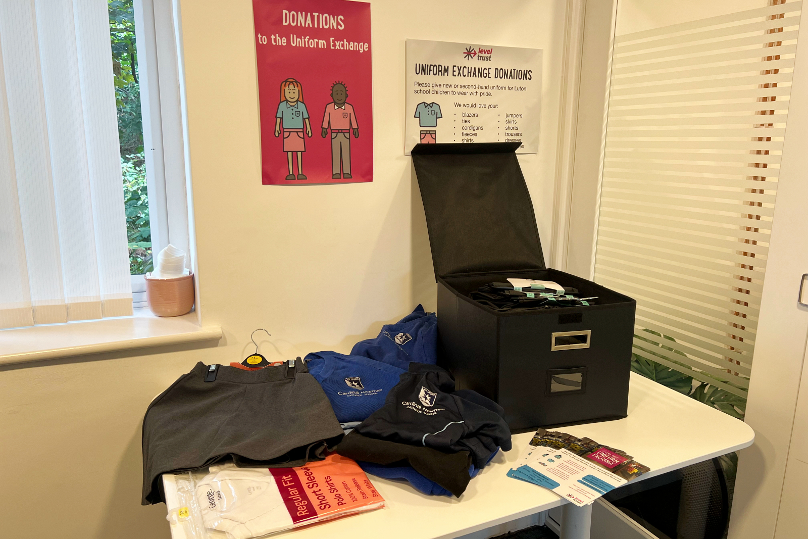 Collection box filled with school uniform items plus further uniform pieces on a table. Leaflets and posters about The Level Trust are also featured in the image.