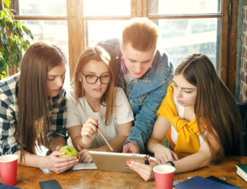 Four young people sat at a table, looking at a laptop with the suggestion they are having a discussion.